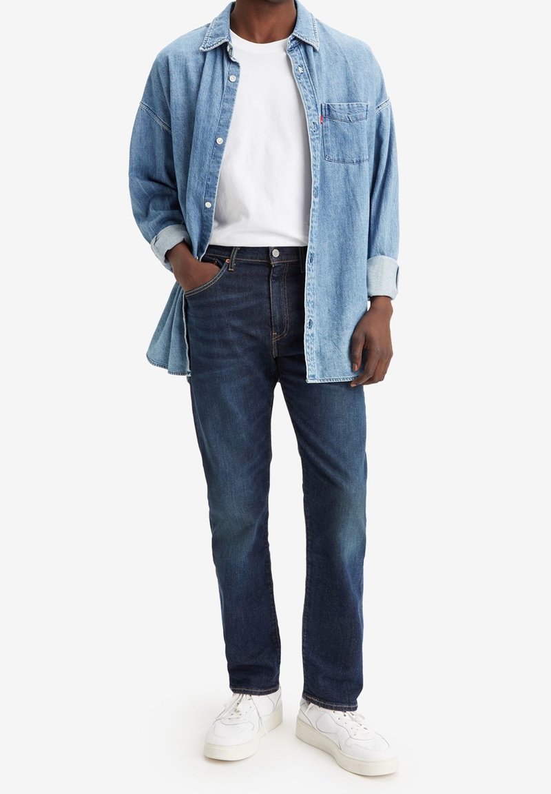 Camisa de mezclilla azul claro, camiseta blanca, jeans azul oscuro y zapatillas blancas. La camisa tiene un bolsillo en el pecho y un diseño de botones.