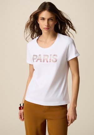 Kvinde med langt brunt hår iført hvid T-shirt med "PARIS" trykt og brune bukser, stående mod beige baggrund.