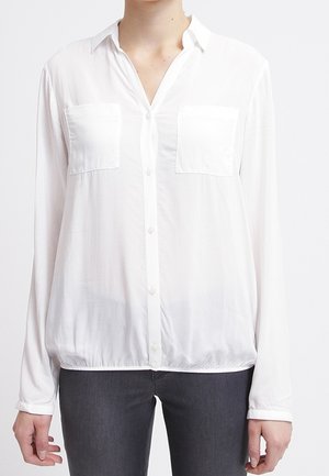 Camicia bianca a maniche lunghe con bottoni, due taschini sul petto, tessuto morbido, colletto e orlo elasticizzato. Indossata con jeans grigio scuro.