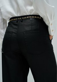 Los pantalones de mezclilla negra presentan un diseño de cintura alta con dos bolsillos traseros y detalles de costura sutiles; un cinturón de cadena dorada realza la cintura.