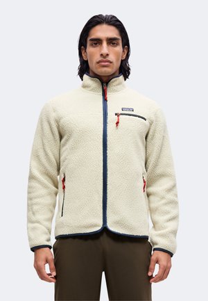 Mann mit dunklen Haaren, der eine cremefarbene Fleecejacke mit marineblauem Besatz und roten Reißverschlussschiebern trägt und vor einem einfachen hellen Hintergrund steht.