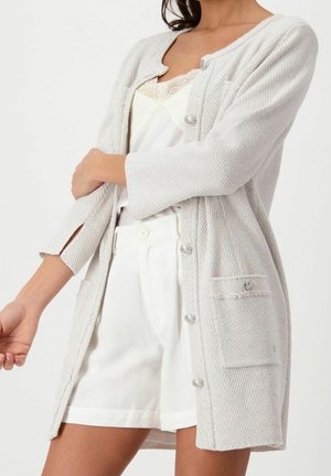 Žena v lehkém šedém texturovaném svetru přes bílý krajkový top a bílé vysoké kraťasy, stojící se založenýma rukama.