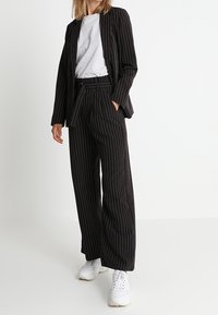 Costume noir à fines rayures avec une coupe ample, comprenant un blazer et un pantalon à jambes larges, associé à un t-shirt gris et des baskets blanches.