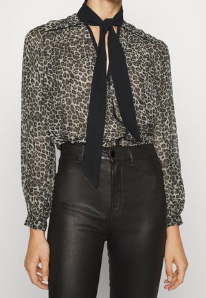 Persoon draagt een doorschijnende blouse met luipaardprint, een zwarte stropdas en hooggetailleerde zwarte leren broek, handen ontspannen langs de zijkant.