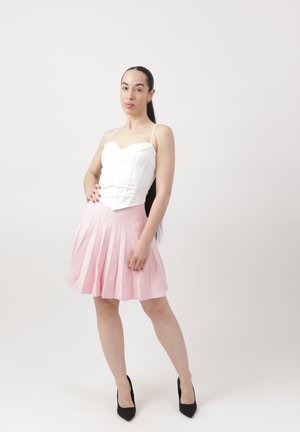 Mujer con cabello largo y oscuro, que lleva una blusa blanca sin mangas, falda plisada rosa y tacones altos negros, de pie frente a un fondo claro y sencillo.