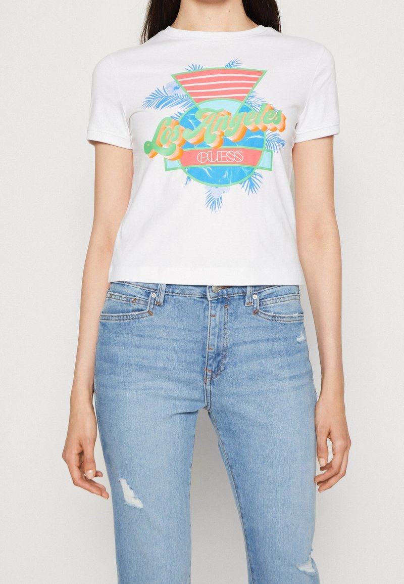 T-shirt en coton blanc avec une impression graphique colorée présentant des palmiers, des rayures, et le texte "Los Angeles Guess." Associé à un jean bleu clair.