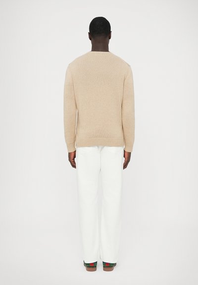 Béžový pletený sveter s rebrovanou textúrou, okrúhlym výstrihom a priliehavými manžetami, v kombinácii s bielymi nohavicami a farebnými teniskami. Pohľad zozadu.
