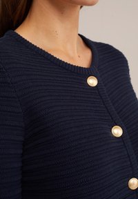 Granatowy kardigan z teksturowanego splotu z okrągłym dekoltem, ozdobiony trzema złotymi guzikami z przodu.