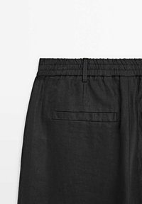 Vue arrière d’un pantalon noir avec taille élastique, passants de ceinture et une poche passepoilée simple sur fond blanc uni.