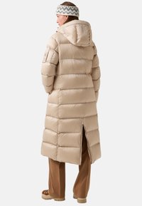 Beige pufferjack met een capuchon, met verticale stiksels, zijopeningen en een ontspannen pasvorm. Gecombineerd met een bruine broek en beige schoenen.