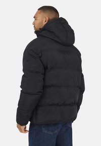 Doudoune noire avec capuche, présentant un design matelassé et des poignets élastiques. Fabriquée en tissu léger et lisse avec une finition mate.