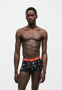 Modèle masculin musclé portant un boxer noir avec des imprimés blancs sur le thème de l'espace et une ceinture rouge portant l'inscription "HUGO", se tenant devant un fond uni.