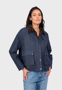 Námořnická modrá voskovaná bunda s hnědým kordovým límcem, vybavená dvěma předními kapsami a patentkami, navržená s uvolněným střihem.
