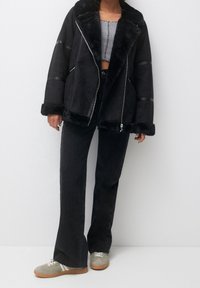 Veste en fausse fourrure noire avec grand col, fermeture éclair et poches latérales, associée à un pantalon en denim foncé évasé et des baskets vertes.