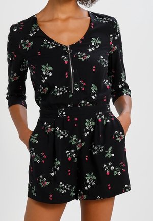 Kobieta ubrana w czarny kombinezon w kwiaty z rękawami do łokcia, zamkiem z przodu i dłońmi w kieszeniach na jednolitym jasnym tle.