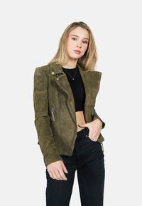 Olijfgroene suède motorjack met een asymmetrische rits, ritszakken en een getailleerd silhouet. Gecombineerd met zwarte high-waisted jeans.