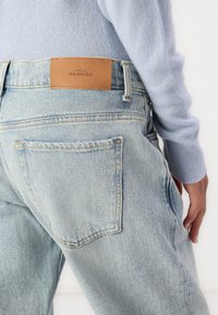 Light blue denim jeans back view with brown leather brand patch, worn with light blue long sleeve sweater and a visible hand resting on the hip.