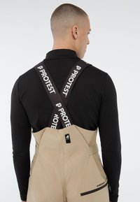 Beige bretels met verstelbare bruine PROTEST banden, voorzien van zijzakken en een gladde textuur. Draagt over een zwart lange mouwen top.