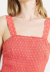 Korálová top s nařasenými detaily a nastavitelnými popruhy s květinovým vzorem. Má texturovaný povrch a přiléhavý korzet s volantovými okraji.