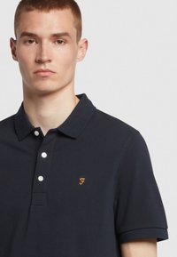 Granatowa koszulka polo z teksturowanej tkaniny, z trzema guzikami z przodu, krótkimi rękawami i małym pomarańczowym logo na piersi.