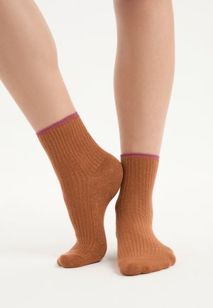 Paire de pieds portant des chaussettes courtes côtelées couleur rouille avec une bordure rose, posés avec un talon levé et les orteils pointés vers le bas.