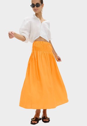 Kvinde iført sorte solbriller, hvid kort bluse, orange rynket maxi-nederdel, guldringe og sorte slip-on sandaler med guldbeklædning.