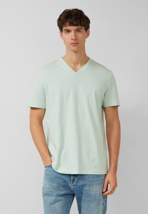 Jeune homme aux cheveux courts et bouclés, portant un T-shirt vert clair à col en V et un jean bleu, debout avec une main dans la poche.