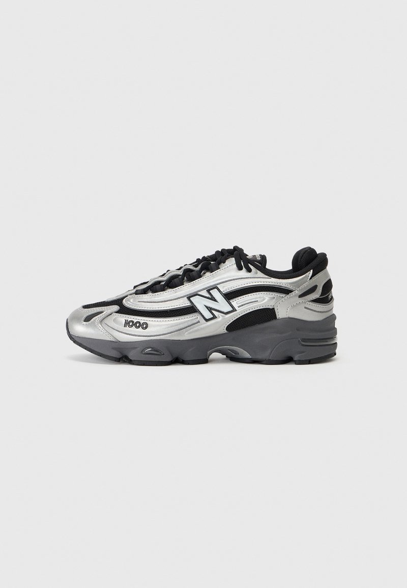 Chaussure de sport New Balance argent et noire avec lacets noirs et semelle épaisse texturée, vue de côté sur un fond gris.