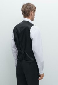 Schwarze Weste aus glattem Stoff mit einem verstellbaren Rückenschnalle, getragen über einem weißen Hemd, mit einem schlanken, maßgeschneiderten Schnitt.