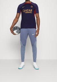 Conjunto deportivo para hombres que consiste en una camiseta de manga corta en navy y rojo con letras doradas, combinada con leggins grises con estampado y zapatillas blancas.