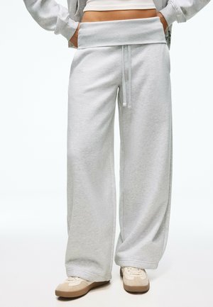 Personne portant un pantalon de jogging gris clair à jambes larges avec taille à cordon et baskets beiges, mains dans les poches, cadré à la taille.