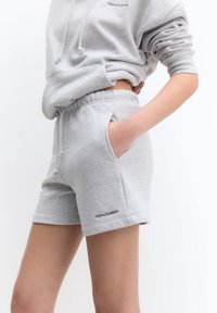 Person med lys grå hættetrøje og matchende shorts med hånden i lommen foran en hvid baggrund.