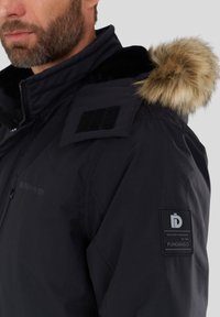 Man in een zwarte winterjas met een bontomrand kap, schouderpatches en een Fundango-logo op de linker mouw.