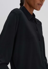 Chemise noire à boutons, confectionnée en tissu lisse, dotée de manches longues, d'un col classique et de boutons sur le devant.