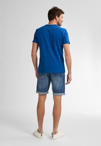 Modrá bavlněná tričko s krátkými rukávy, spárované s ohrnutými džínovými šortkami. Model má na sobě světlé tenisky. Jednoduché pozadí.