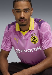 Camiseta de fútbol rosa con acentos morados y cuello amarillo. Presenta los logotipos de "Puma" y "Evonik", y dos estrellas encima de un emblema del club redondo.