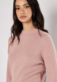 Hellrosa, gerippter Pullover mit hochgeschlossenem Kragen, strukturiertem Stoff und markanten weißen Häkelverzierungen entlang der Schulternähte.