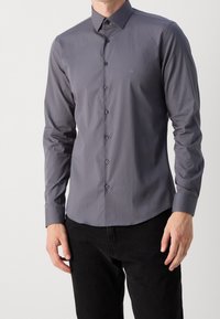 Camisa de manga larga gris hecha de una tela suave con cuello de botón, con un sutil logo en el pecho y botones a juego.