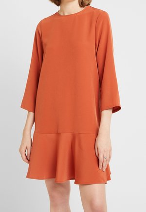 Frau trägt ein locker sitzendes, verbrannt orangefarbenes Kleid mit dreiviertel Ärmeln und ausgestelltem Saum, steht vor einem schlichten, hellen Hintergrund.