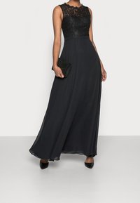 Svart maxi-klänning med spetslivstycke och utan ärmar, flödande chiffonskjol, och matchad med en svart kuvertväska. Elegant silhuett.