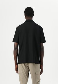 Camisa polo preta de manga curta com colarinho, apresentando um corte limpo, um ajuste ligeiramente solto e uma textura suave, combinada com calças cargo bege.
