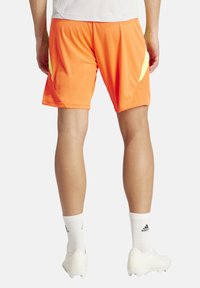 Heldere oranje sportshorts met gele accenten, netachtige textuur en een losse pasvorm, gecombineerd met witte sokken en voetbalschoenen.