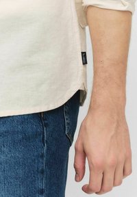 Lichtbeige shirt van zachte stof, met opgerolde mouwen en zijsplitsen. Opvallend zwart label aan de linkerkant. Gekleed met een blauwe jeans.