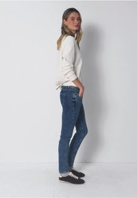 Camiseta blanca de manga larga, jeans ajustados azules y zapatillas negras. La camiseta tiene un corte relajado; los jeans presentan un diseño clásico de cinco bolsillos.