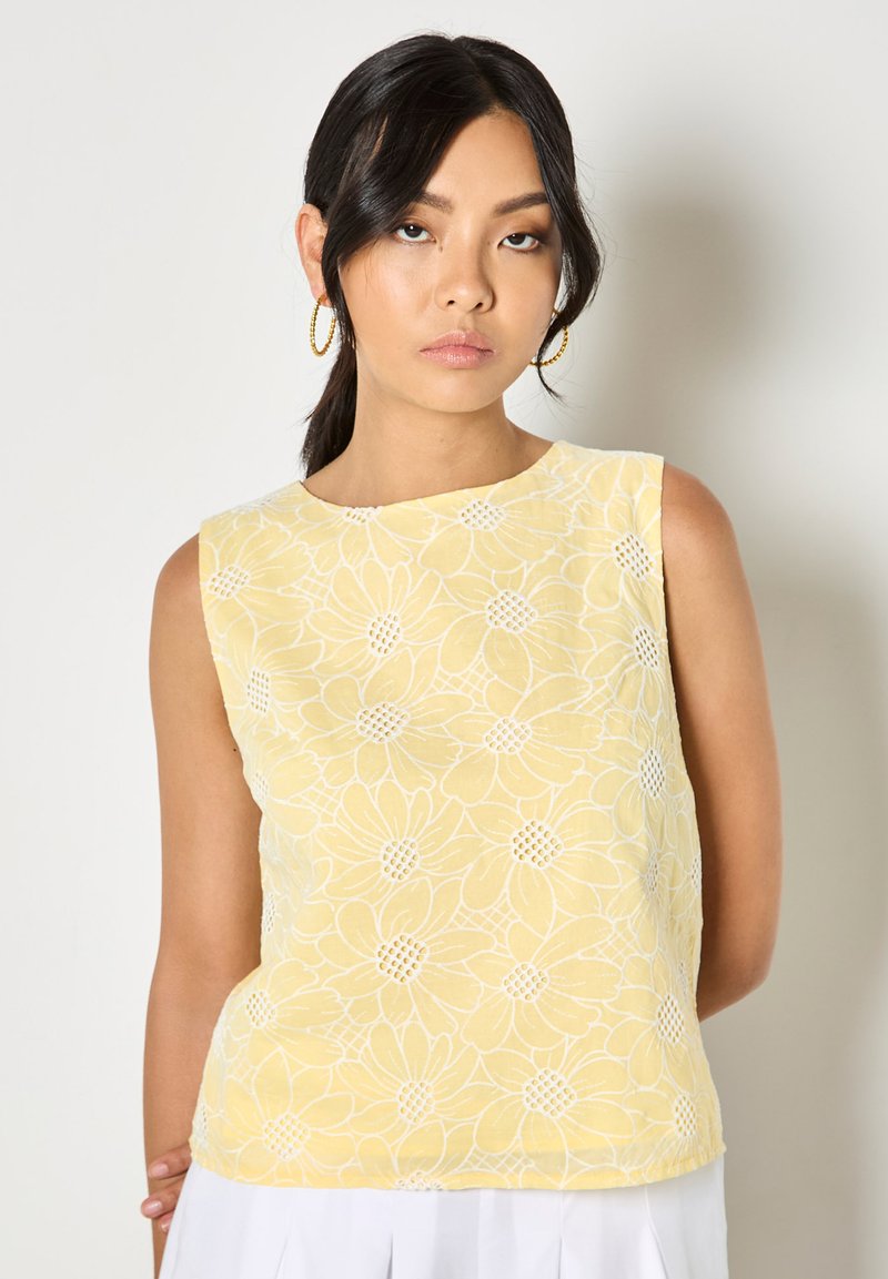 Jeune femme aux cheveux foncés portant des boucles d'oreilles en or en forme d'anneau et un haut sans manches jaune avec des motifs floraux blancs, debout devant un fond clair.