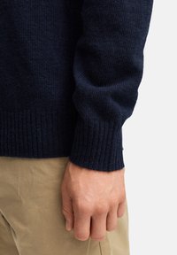 Granatowy sweter dzianinowy z prążkowanymi mankietami. Tekstura wydaje się miękka i ciepła, kontrastując z jasnobeżowymi spodniami noszonymi pod spodem.