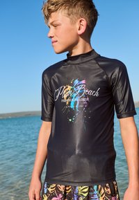 Juodas trumpomis rankovėmis apsaugos nuo saulės marškinėlis su spalvingu purslų raštu ir užrašu „Palm Beach“, derinamas su margintais daugiaspalviais šortais.