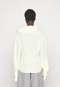 Person med kort hår som bär en långärmad ljusgul blus med volangkrage och ljusgrå byxor, vänd bort.