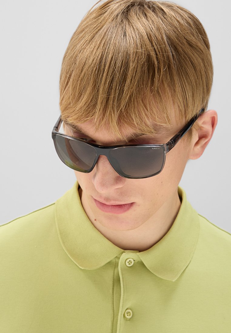 Okulary przeciwsłoneczne z ciemnoszarym oprawkiem i szkłami, o gładkiej fakturze i nowoczesnym kształcie. Noszone z jasnoróżową koszulą z kołnierzykiem.