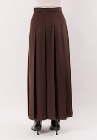 Gonna lunga plissettata marrone abbinata a stivali neri con tacco, vista da dietro su sfondo chiaro e uniforme.